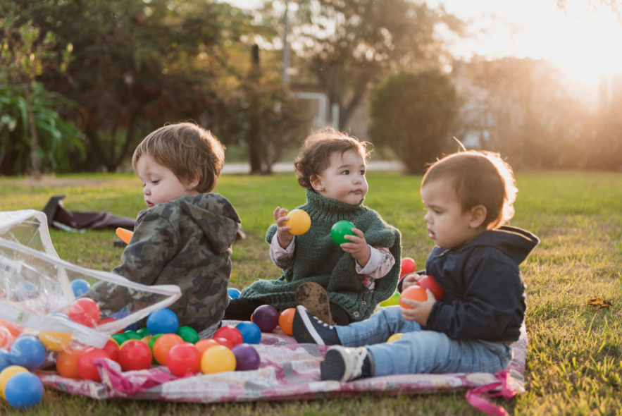troje małych dzieci siedzi na kocu i bawi się w ogrodzie plastikowymi piłkami
